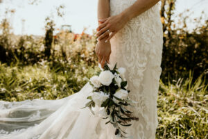 Bride wearing a lace wedding dress holding a white bouquet outdoors.